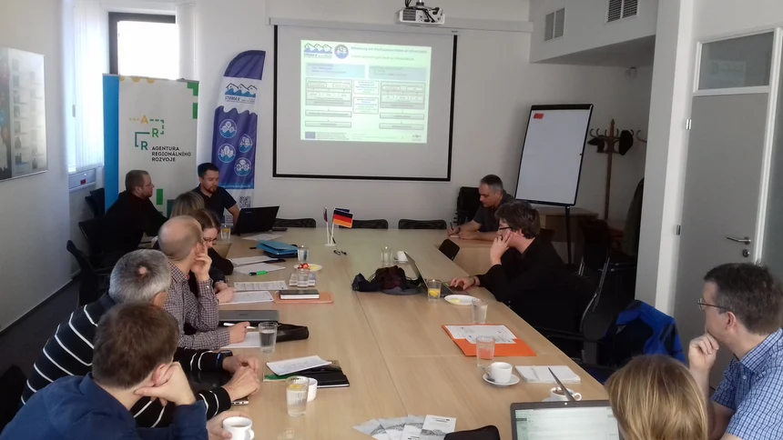 Powerpointová prezentace projektových partnerů, kteří představují rozpracovanost svých pracovních celků, další projektoví partneři sedí u stolu a naslouchají.