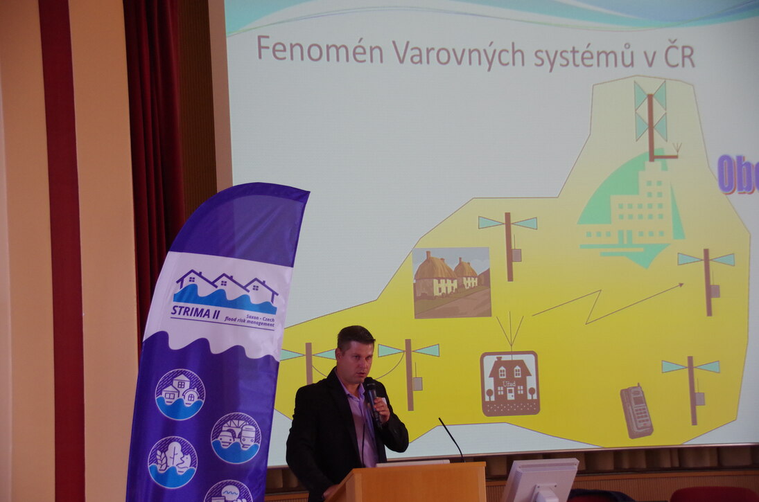 Český přednášející pan Vladimír Pavlík přednáší na téma přeshraniční systém včasného varování. Vedle něj je vidět vlajka projektu STRIMA II.