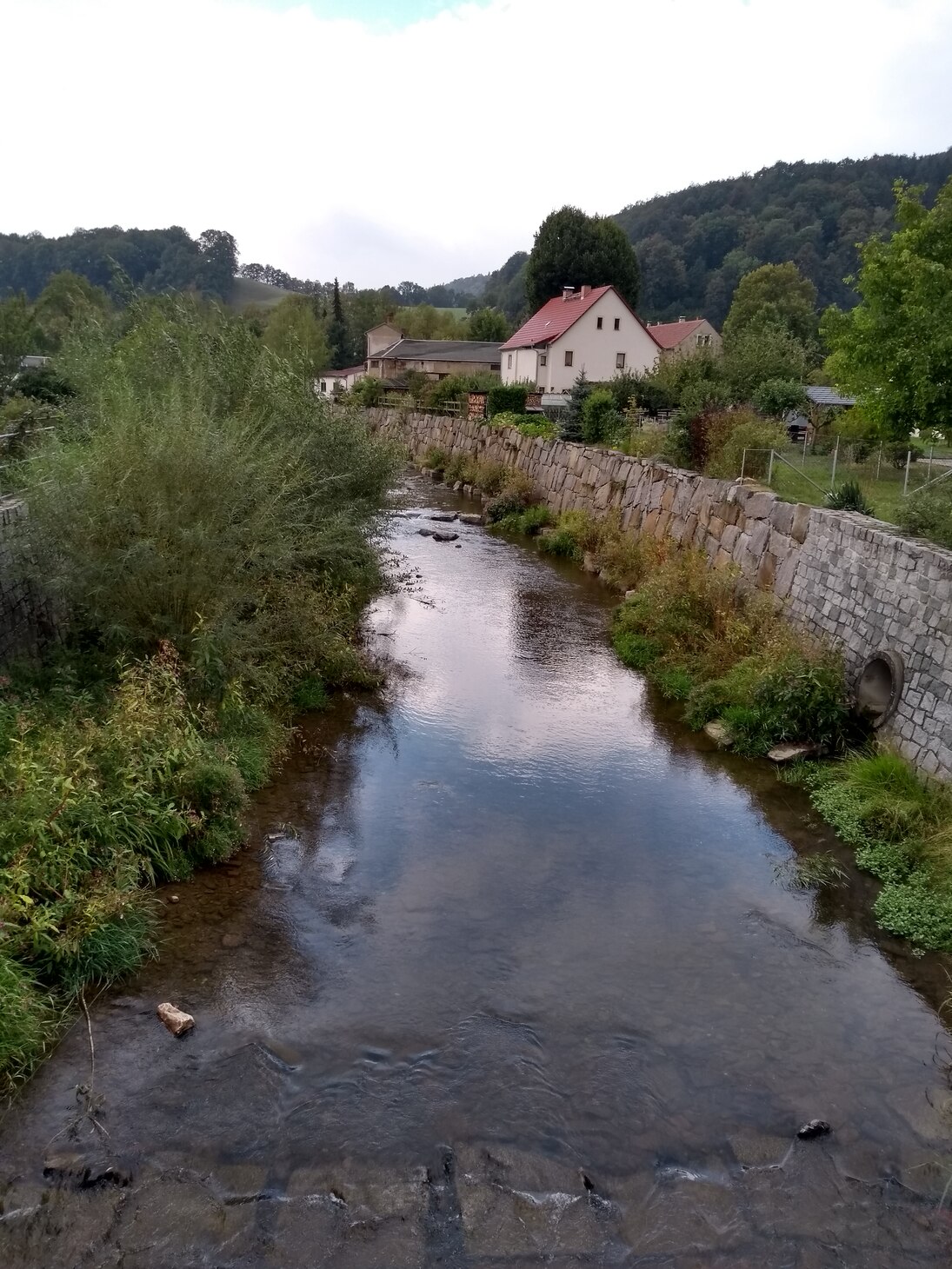 Mit Blick von einer Brücke ist ein Gewässerabschnitt der Müglitz zu sehen. Private Grundstücke sind durch eine Steinschüttungs-Mauer vorm Wasser geschützt. Das Gewässer ist nur wenig gefüllt.