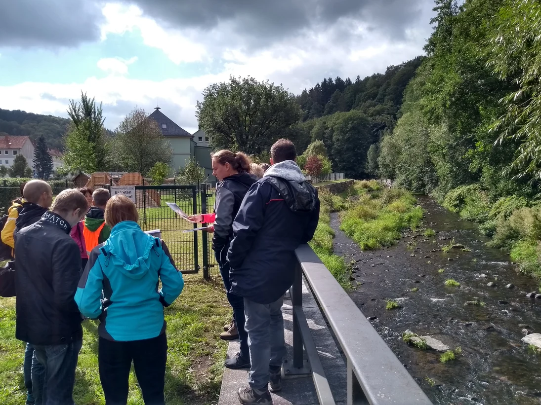 Die Teilnehmer stehen beisammen und erhalten Erläuterungen zum nebenan befindlichen Gewässerabschnitt der Müglitz im Ortsteil Schlottwitz. Im Hintergrund sind Gebäude und die Kita von Schlottwitz ersichtlich.