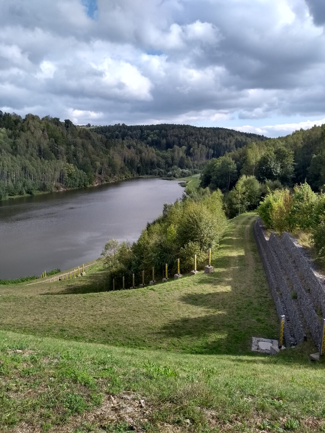 Mit Blick von der Dammkrone ist das Hochwasserrückhaltebecken Lauenstein zu sehen, welches teilweise gefüllt ist. Im Bildvordergrund sind die Pegellatten zur Ermittlung des Wasserstandes zu erkennen. 