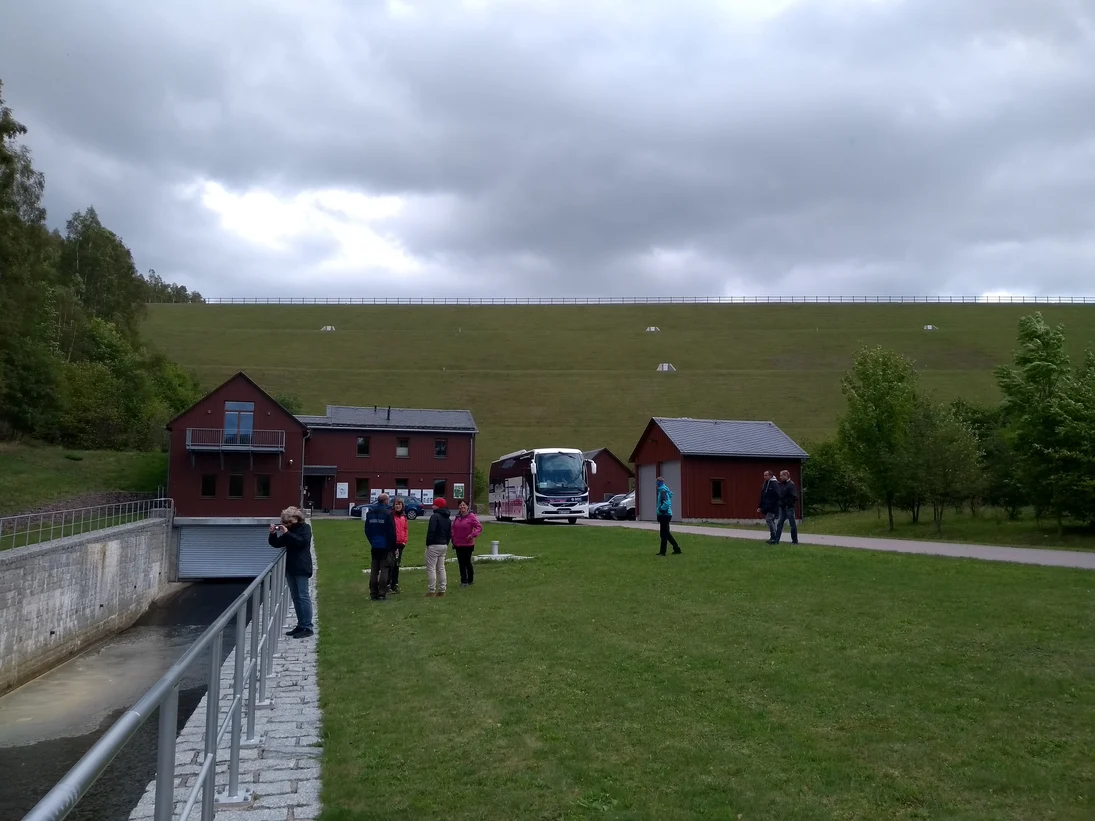 Das Bild wurde vor den Gebäuden der Staumeisterei Müglitz aufgenommen. Im HIntergrund ist der Damm des Hochwasserrückhaltebeckens Lauenstein zu sehen. Einzelne Exkursionsteilnehmer stehen mit dem Staumeister zusammen oder besichtigen die Umgebung.