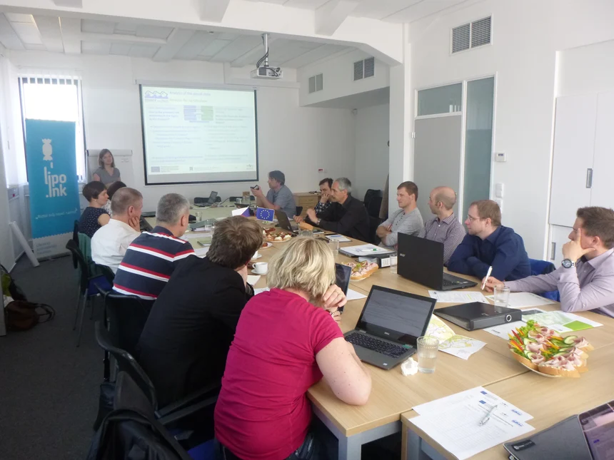 Všichni účastníci projektu sedí u velkého stolu v zasedací místnosti a přihlížejí power-pointové prezentaci